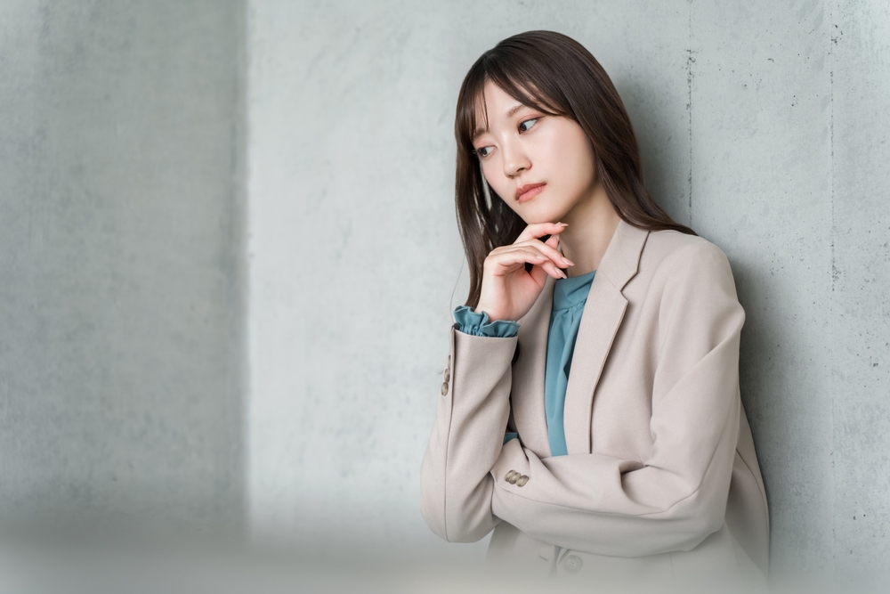 灰色の壁に寄りかかり、あごに手を添えて悩んでいるスーツ姿の女性